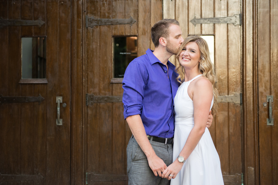 a man kisses his fiancé on the head. She is smiling at the camera wearing a white dress, he is wearing a purple shirt. They are holding hands, and there is an old wooden door behind them. Engagement session at UWO by London wedding photographer Life is Beautiful Photography