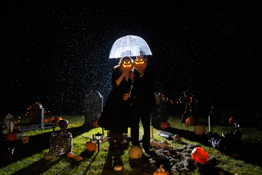 a couple poses at hessenland with jack-o-lanterns in front of their faces, and a lit up cemetary behind them at their halloween wedding