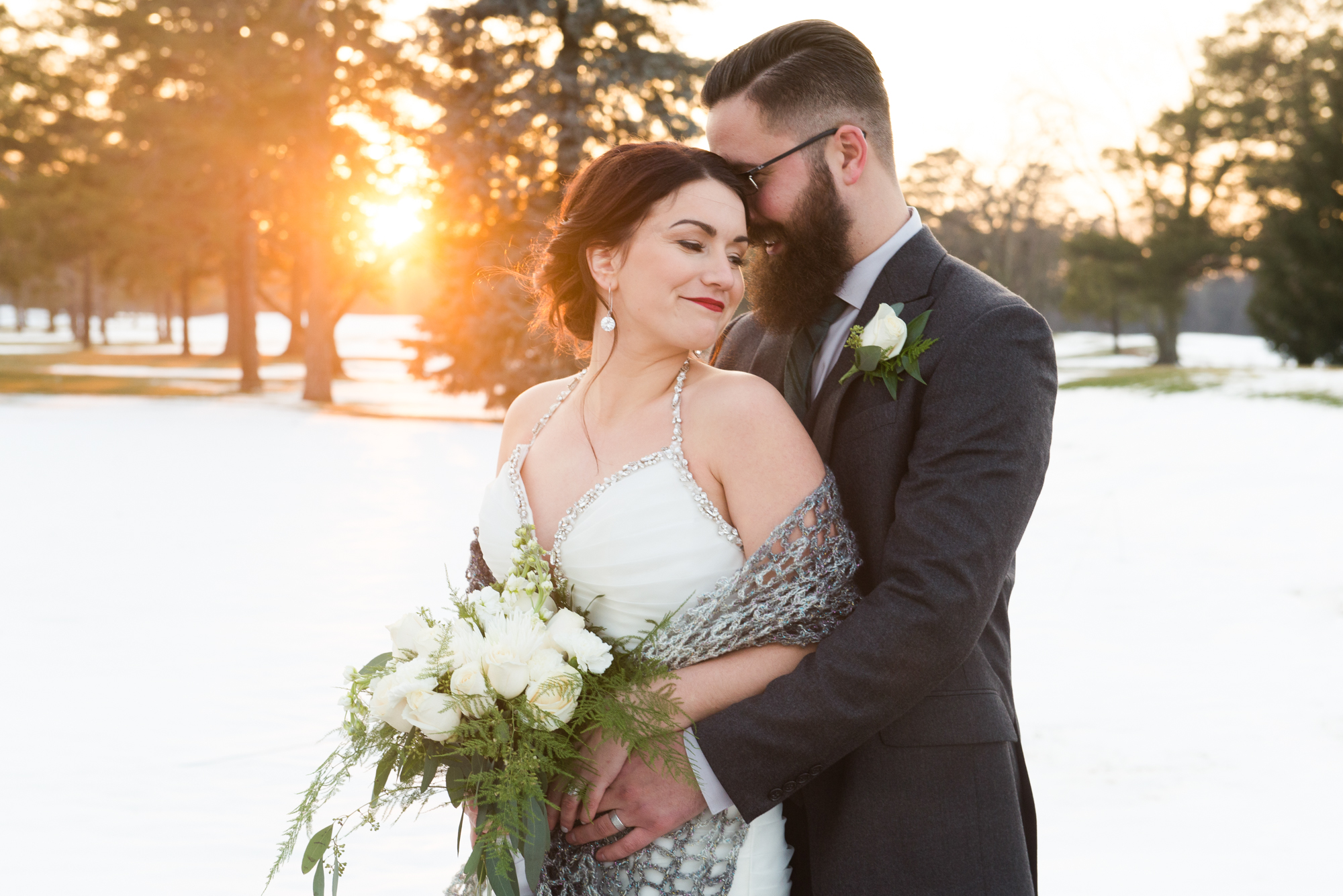a groom stands behind a bride and wraps his arms around her. there is snow in the background and the sun is setting behind them. by toronto wedding photographer life is beautiful photography