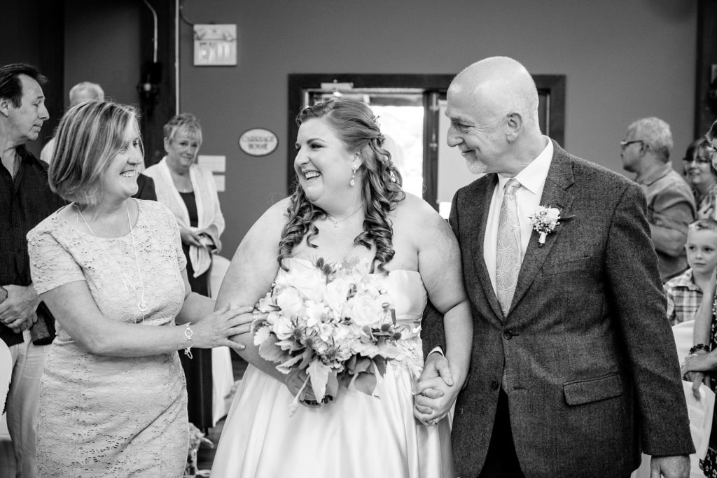 a bride laughs with her parents as they walk her down the aisle
