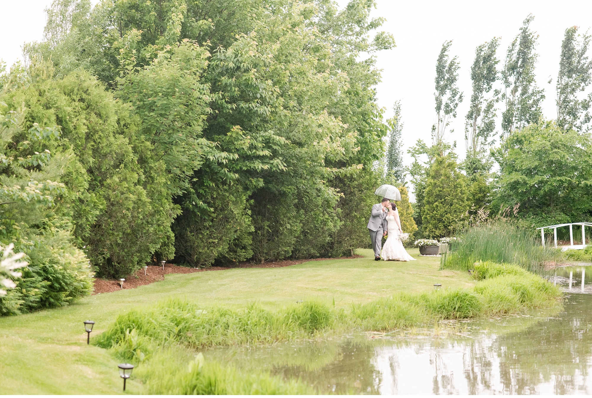 a couple walks across the grass beside a pond on their wedding day. the groom is holding an umbrella above their heads. photo by kelowna wedding photographer life is beautiful photography