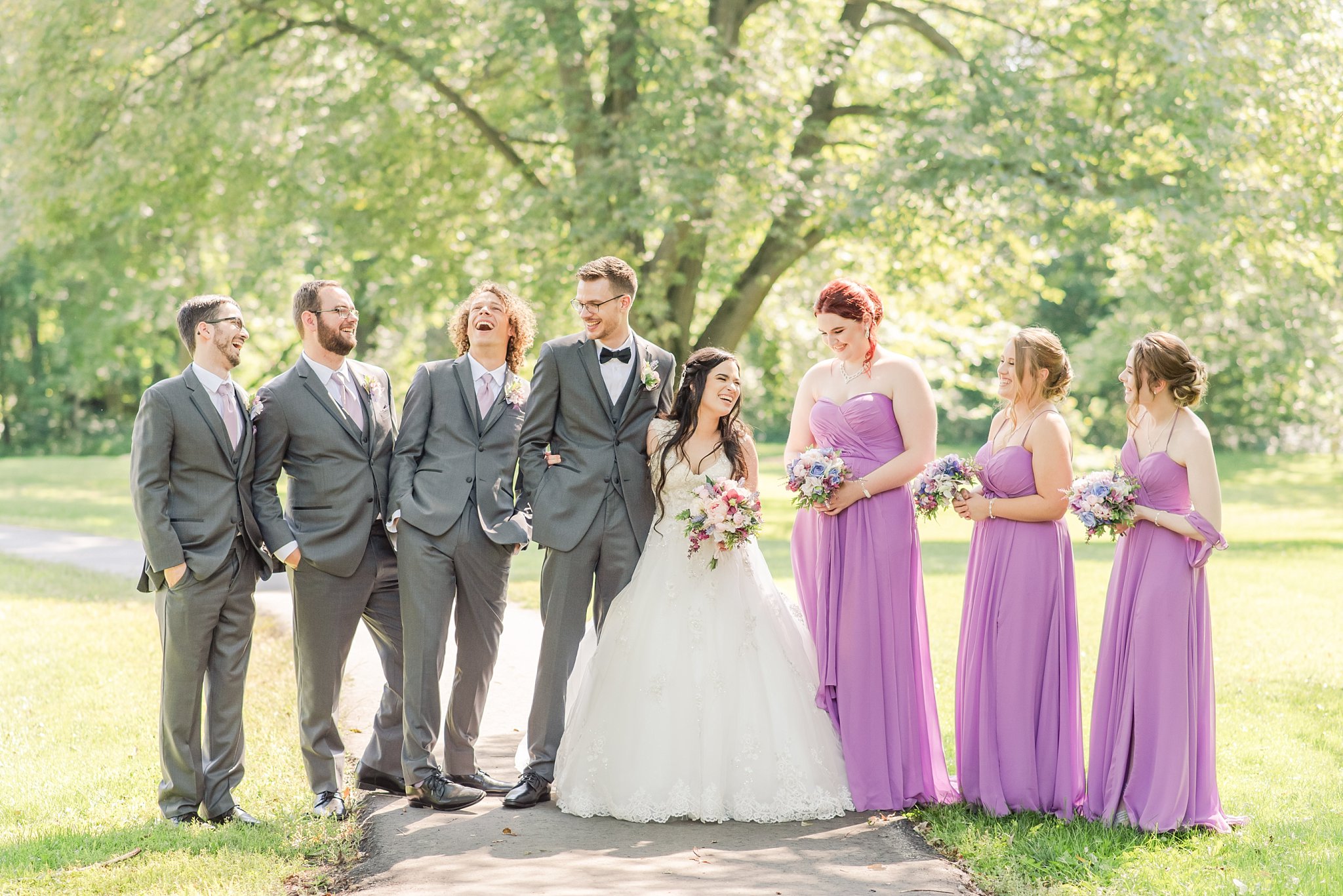 a wedding party stands on a path among trees, laughing with one another