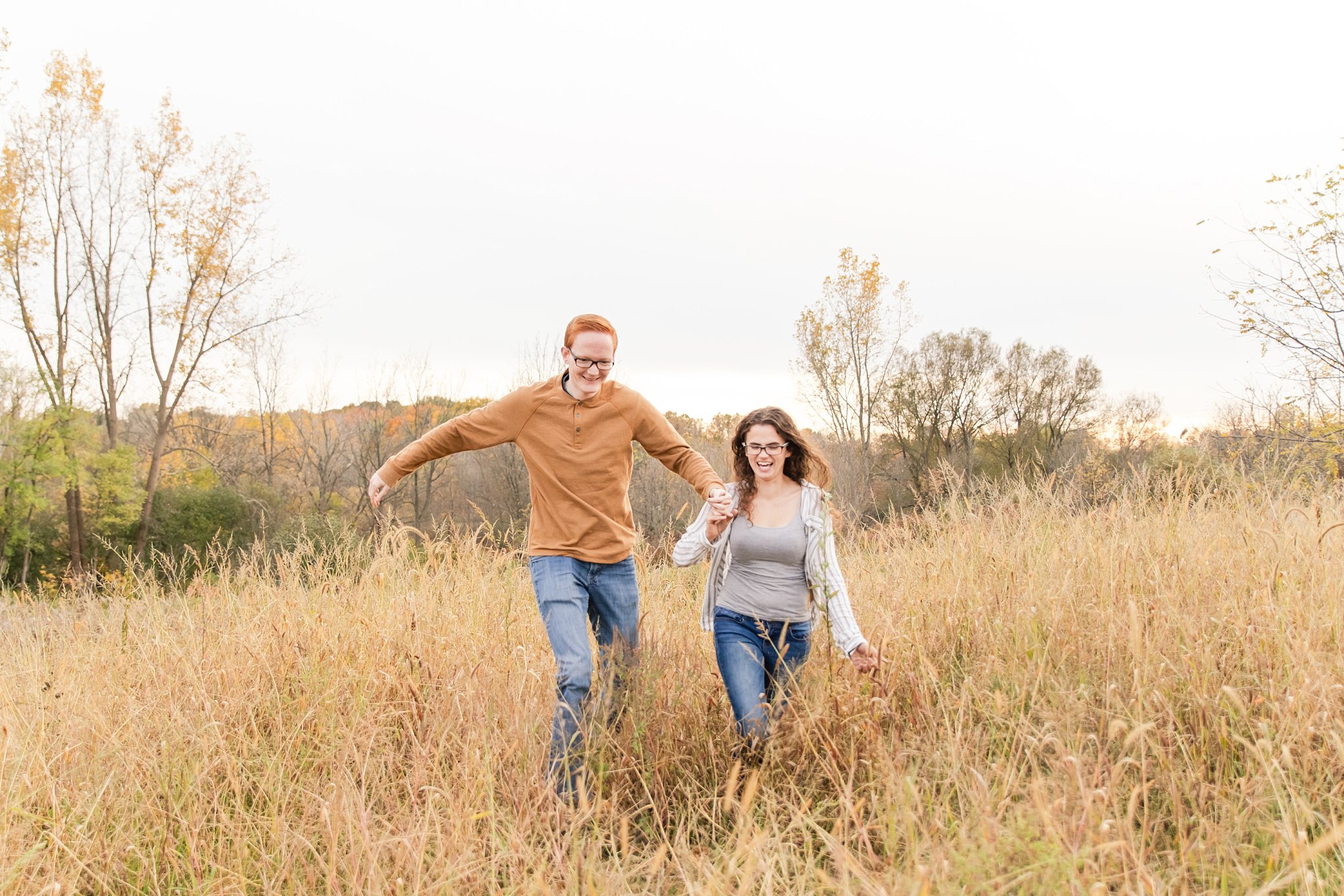 a man and woman laugh and hold hands while running through a field during their engagement session