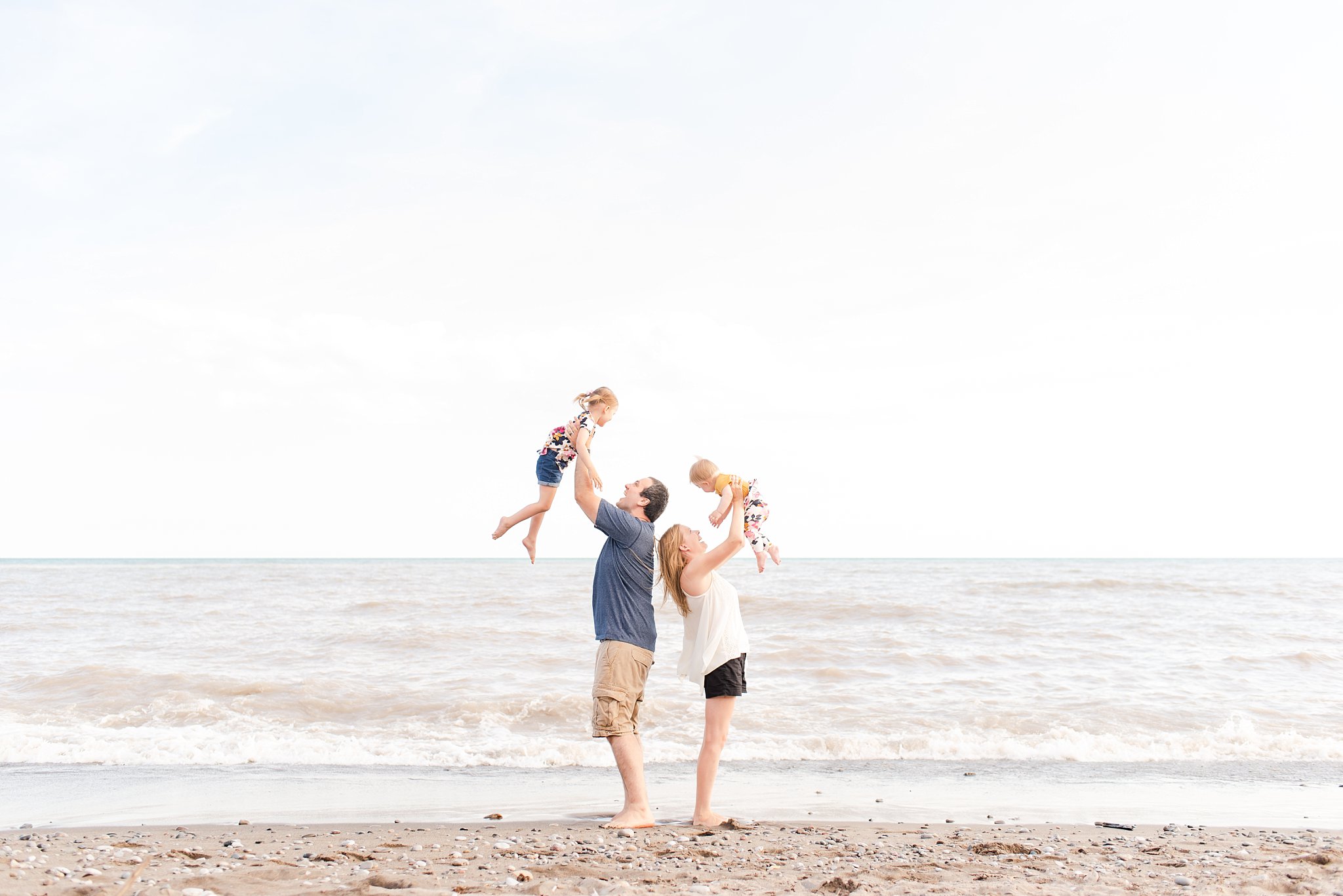family photos in port stanley; mom and dad lift their daughters in the air on the beach with the water in the background