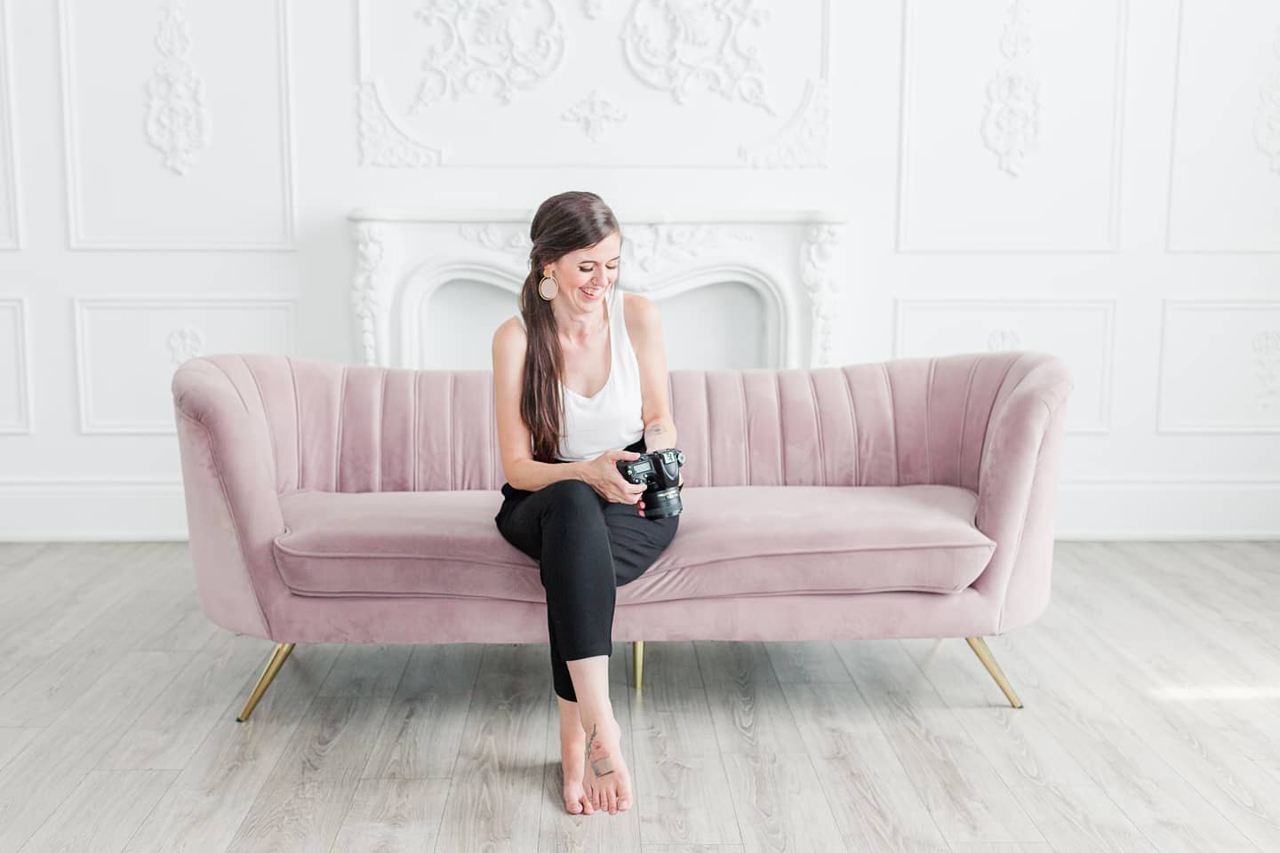 Sandra, Toronto Wedding Photographer and owner of Life is Beautiful Photography, sits on a light pink couch in a room with white walls and light wood floors. She is laughing while looking down at a camera she's holding.