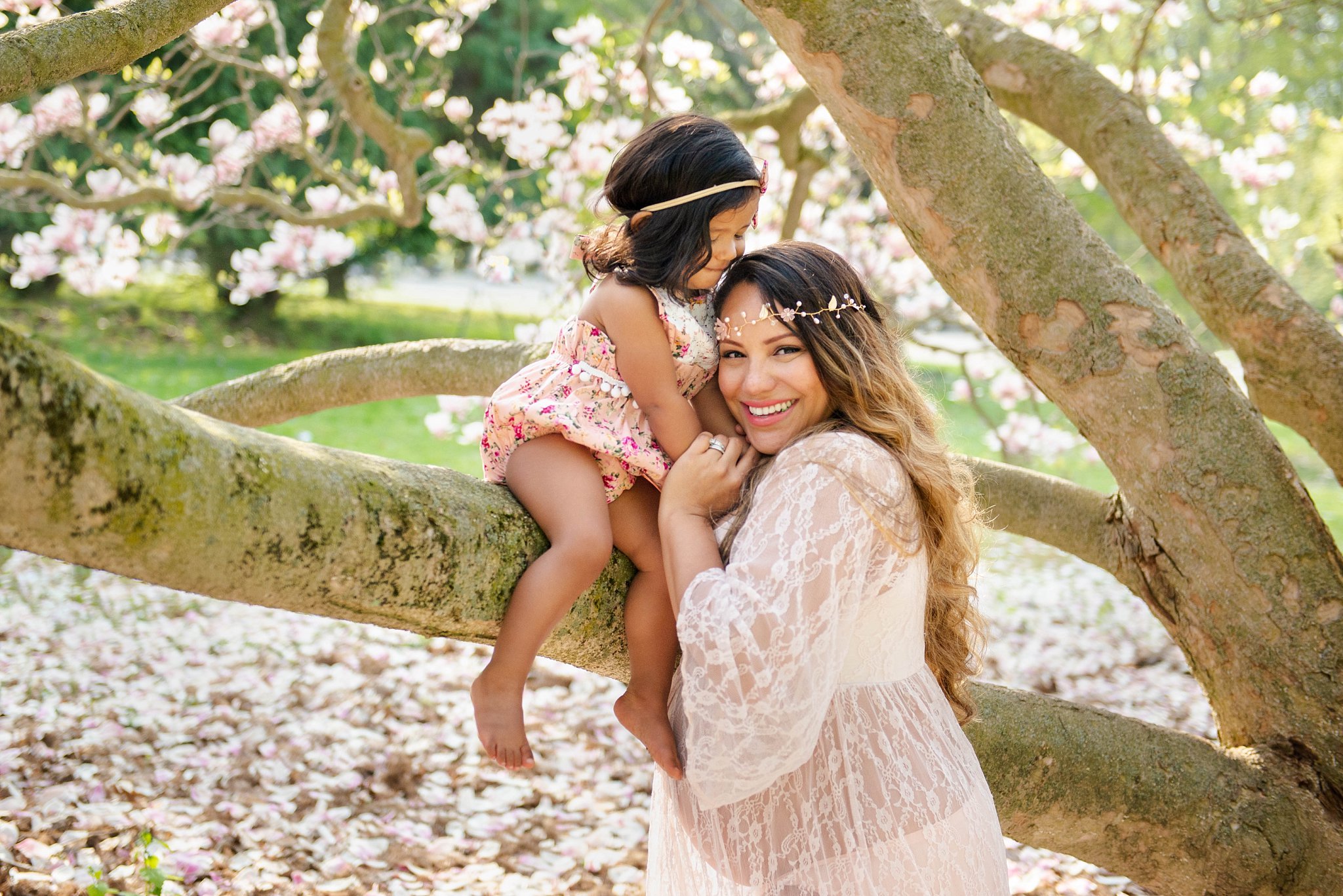 a little girl sits on a magnolia tree branch and holds hands with her mom, who is smiling at the camera