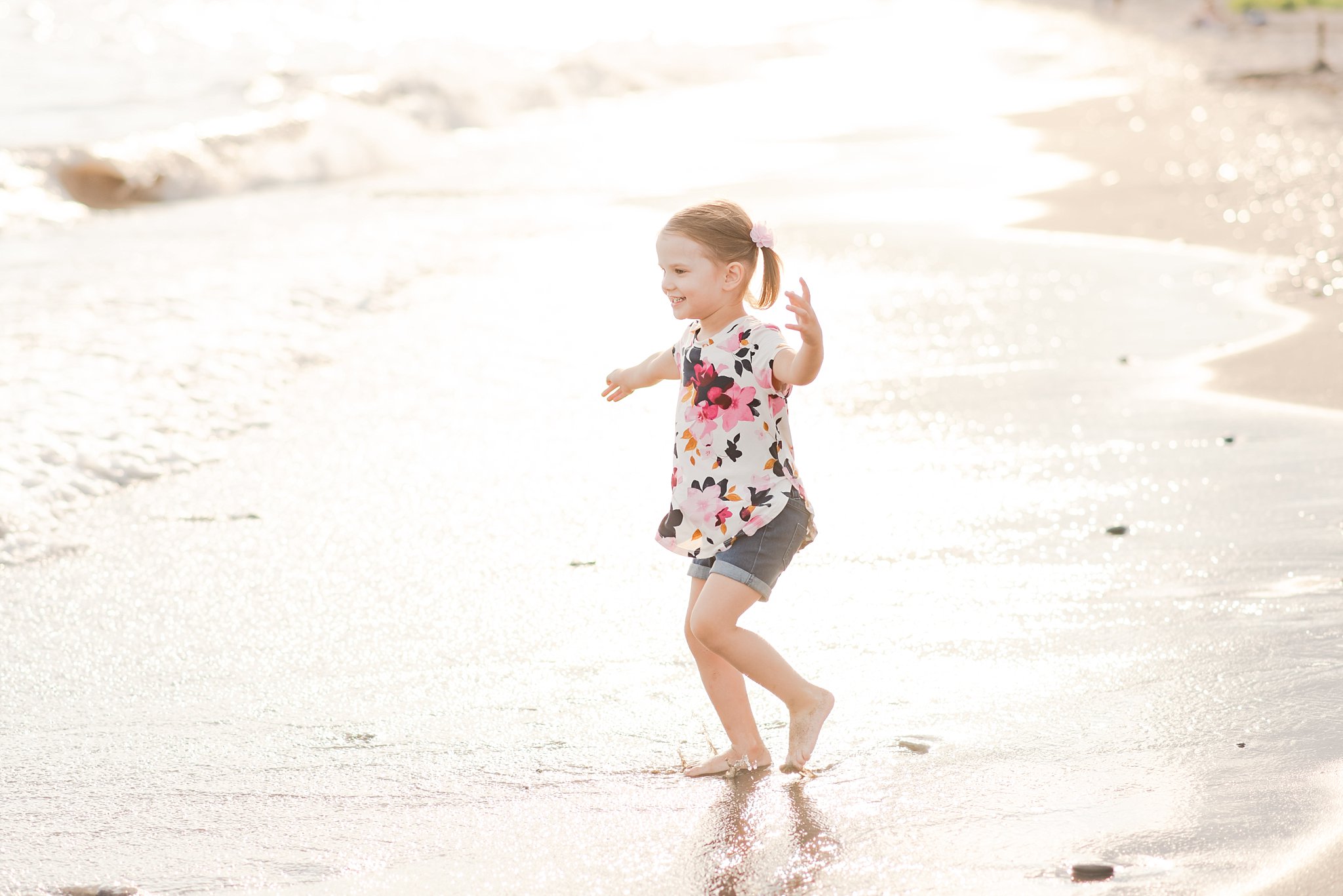 family photography london ontario. a little girl wearing a flowery shirt and blue shorts splashes in water on the shore of Port Stanley
