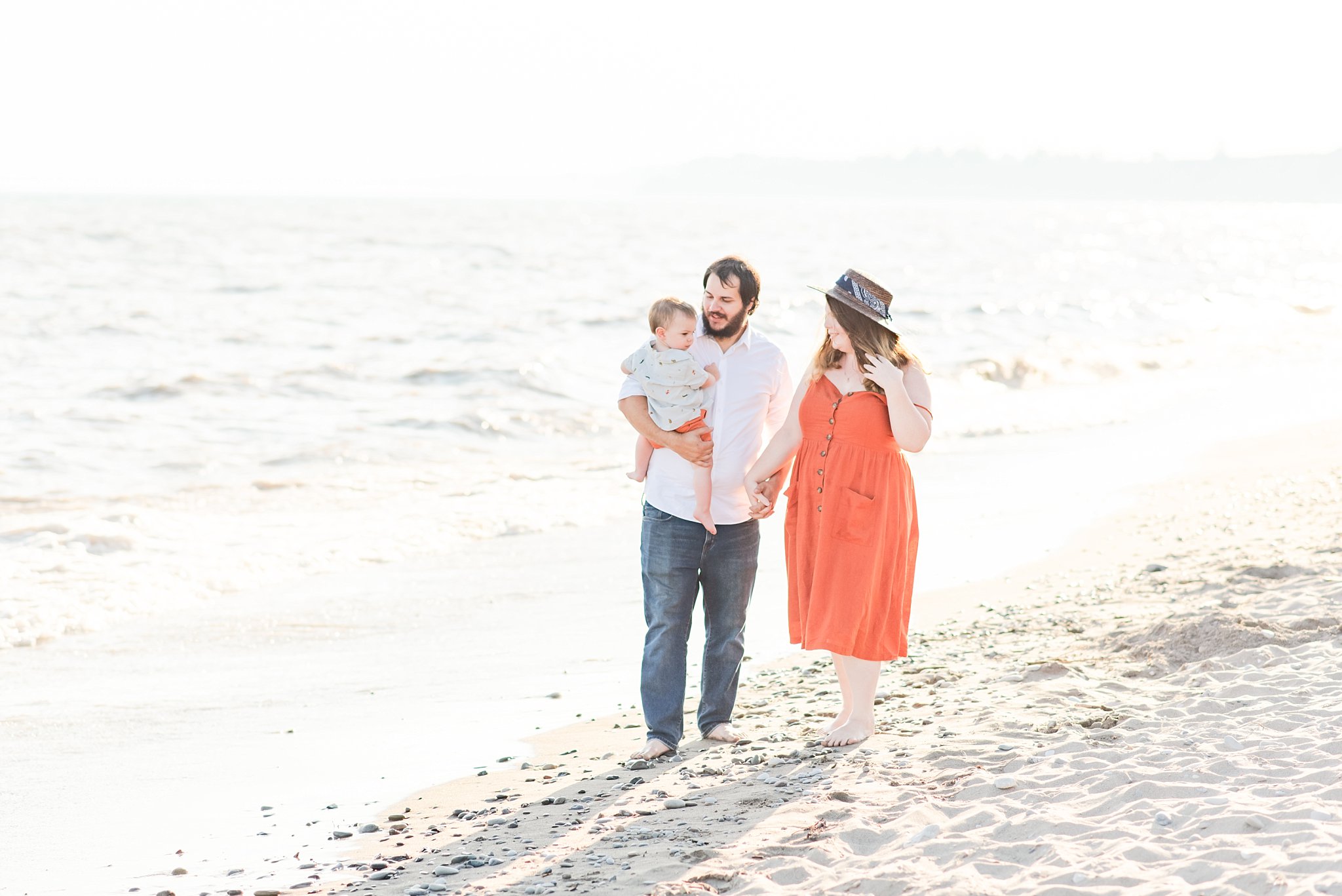 a woman in an orange dress, wearing a hat, holds hands with a man in a white shirt and blue jeans. the man holds a blonde boy wearing a light blue shirt and orange shorts. they're walking down the beach near the water at sunset. family photography london ontario