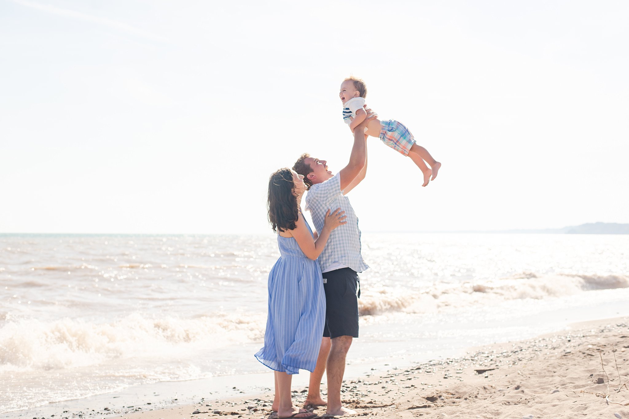 family photography london ontario. a woman in a blue dress stands behind a man in a white and blue shirt and blue shorts. he is lifting their son high into the air. the boy is wearing a white shirt and multicoloured plaid pants. there is sun shining off waves splashing behind them.