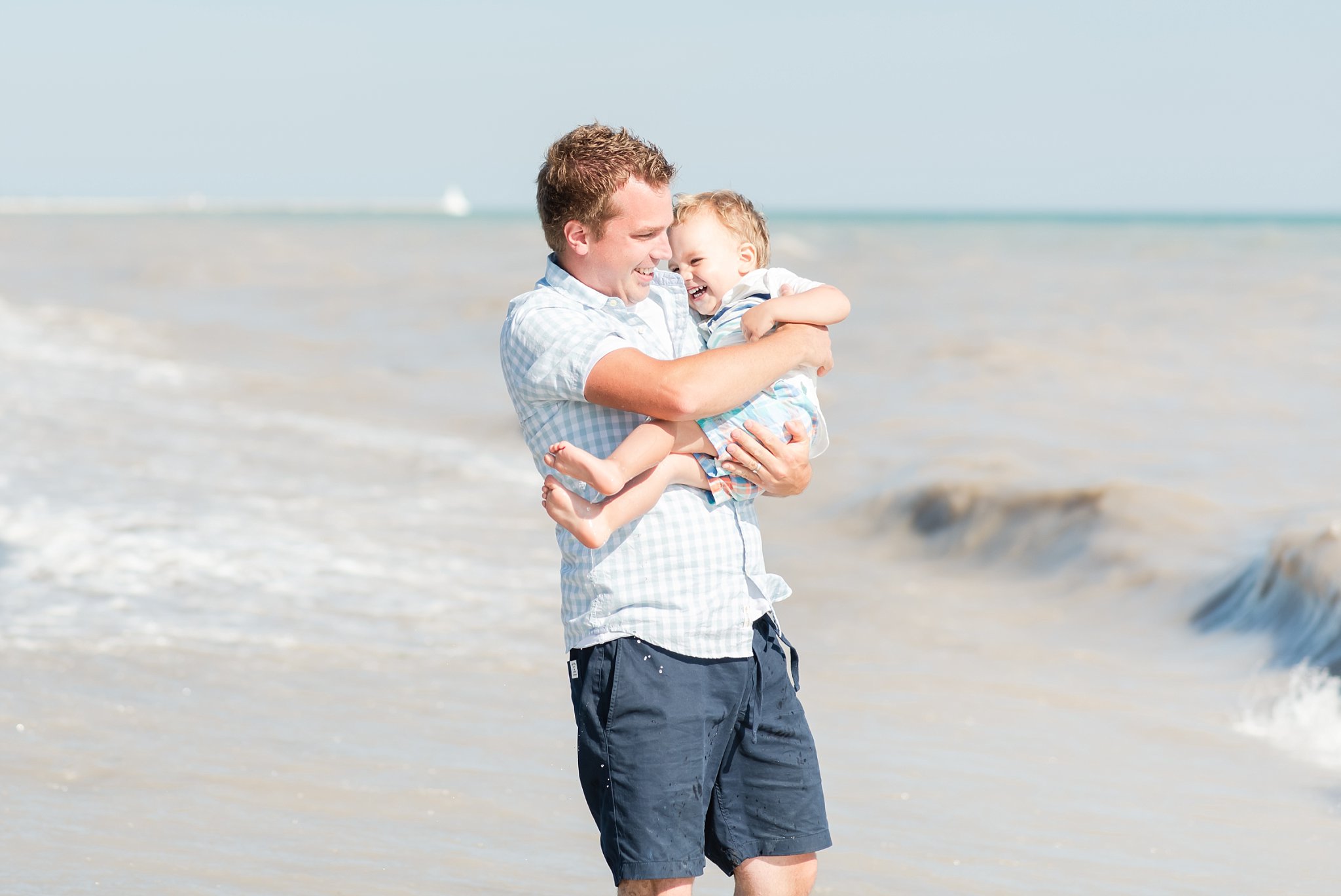 a dad holds his son in the air before leaning in to splash the boy's feet in the waves in port stanley. family photography london ontario. by life is beautiful photography