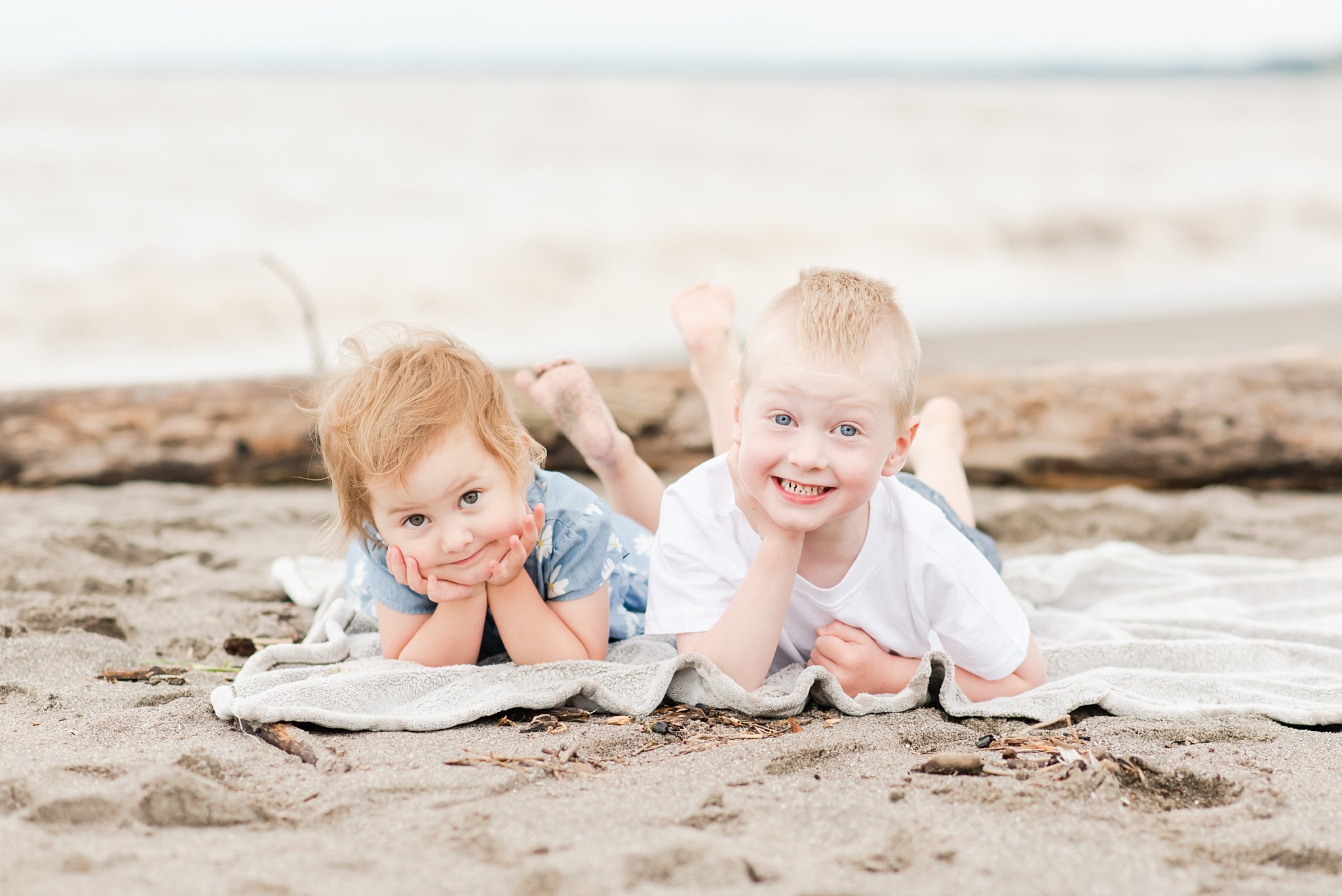 family photography london ontario. a little boy and a little girl lay on their stomachs with their chins in their hands, on a blanket at the beach. they're both blonde, he is wearing a white shirt and she is wearing a blue and white dress.
