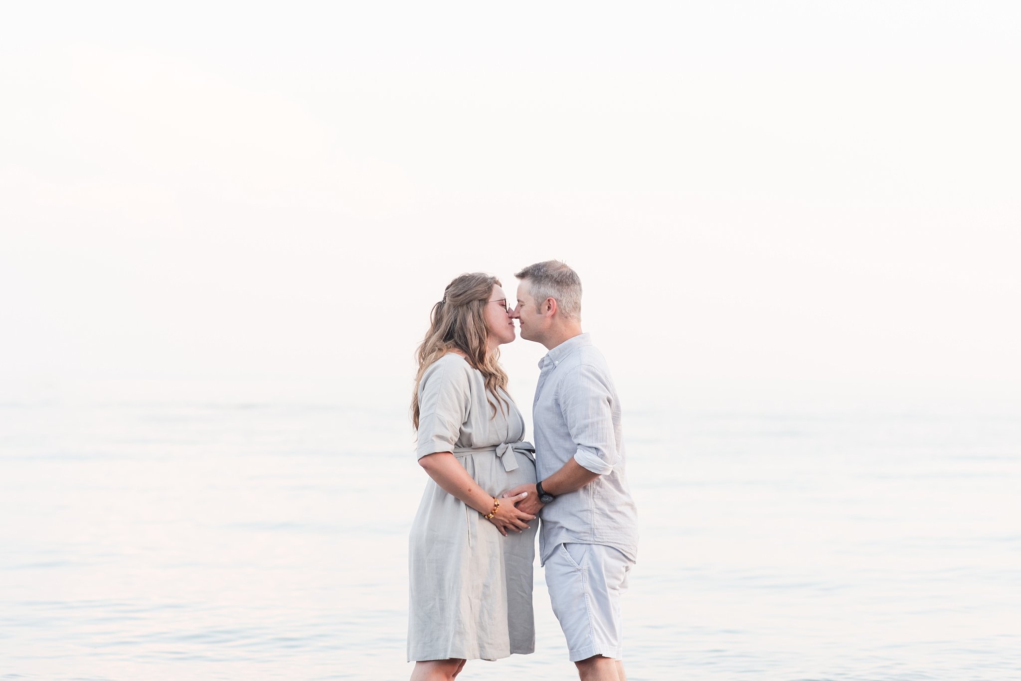 a pregnant woman in a grey dress leans in to kiss her husband, who is wearing a light grey shirt and white shorts. she has long blonde hair and he has grey hair. their hands are holding the woman's belly for their maternity session. family photography london ontario.