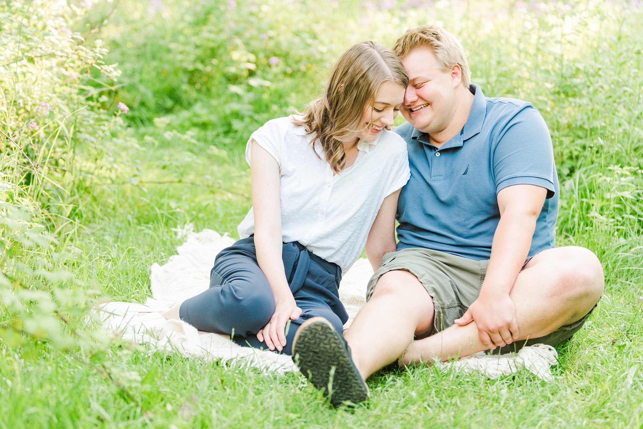 a woman leans her head in towards her fiancé while they laugh together. they're sitting on a blanket in a field during their engagement session with wedding photographers in london ontario life is beautiful photography