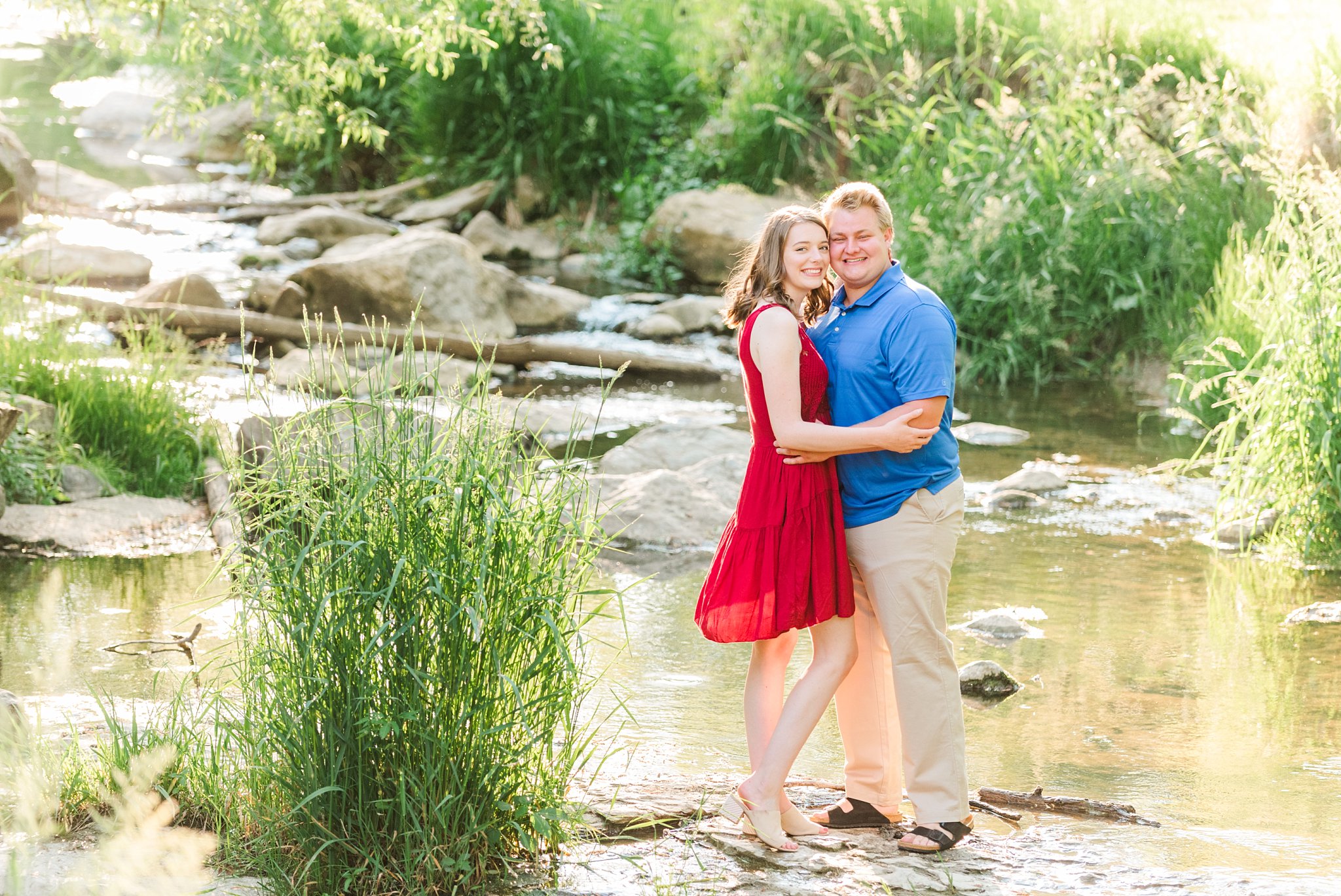 a couple stands on a rock in a creek with their arms wrapped around one another