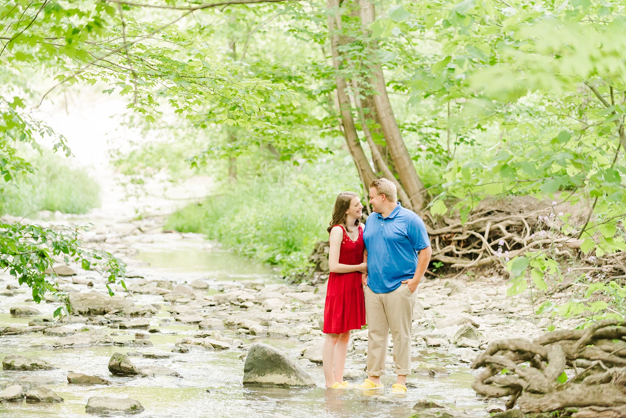 a couple stands along the edge of a creek and smile at one another