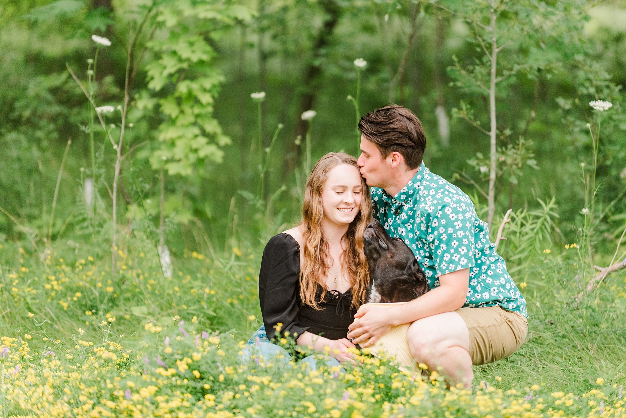 a man kisses a woman's head as they sit in a meadow during their engagement session at guelph arboretum