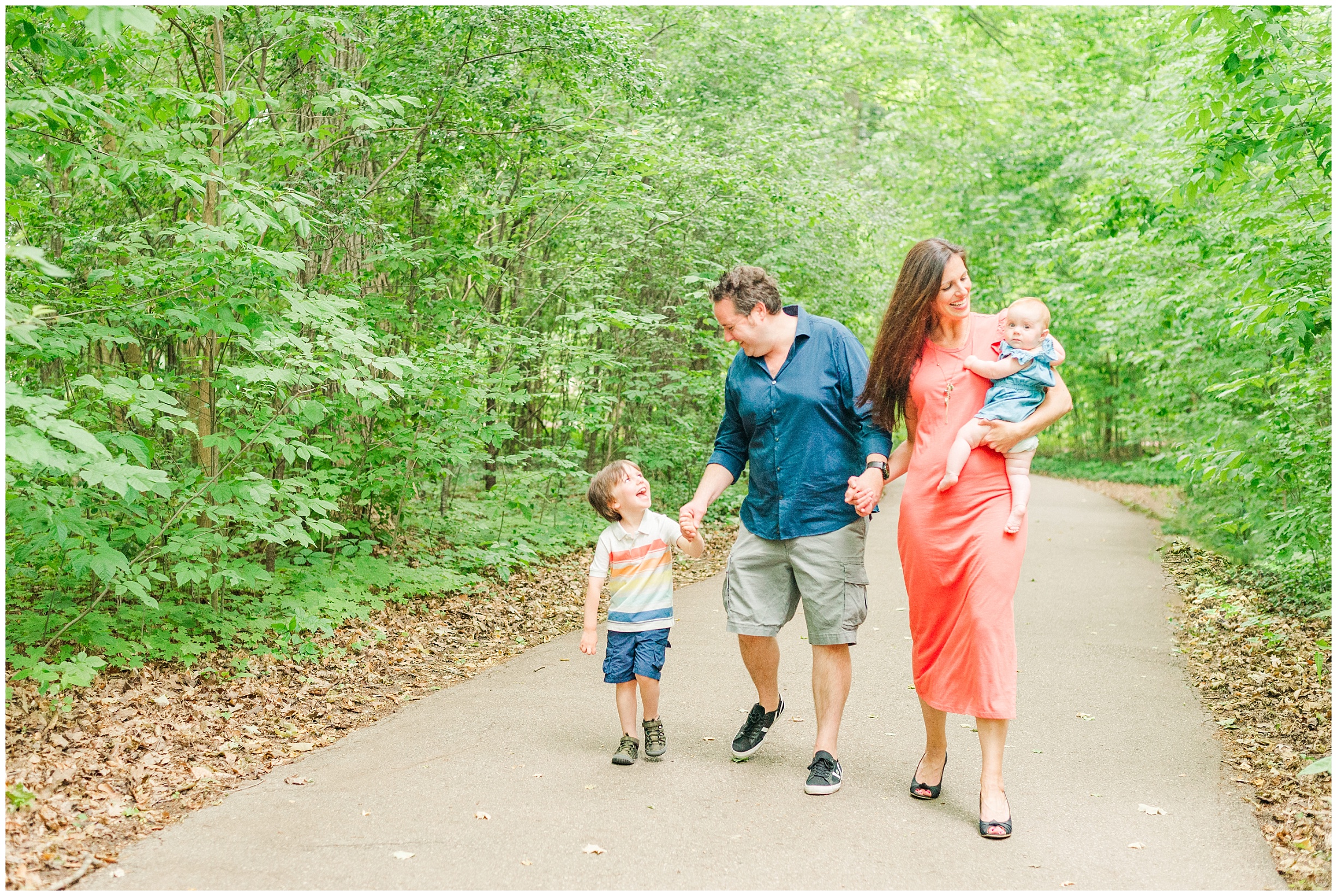A family of four walks down a paved path surrounded by trees. A little boy is holding his dad's hand and looking up at him, dad is looking down at his son. Mom is holding their daughter on her hip and smiling while looking at her.