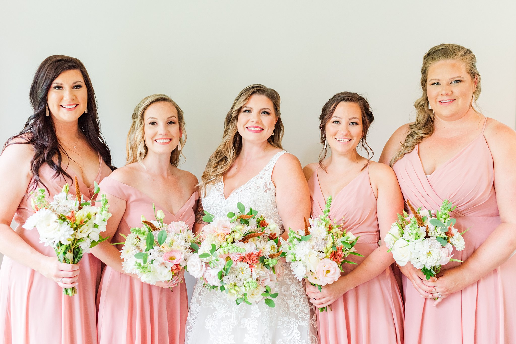 a bride and four bridesmaids wearing pink dresses, all hold bouquets and smile at the camera