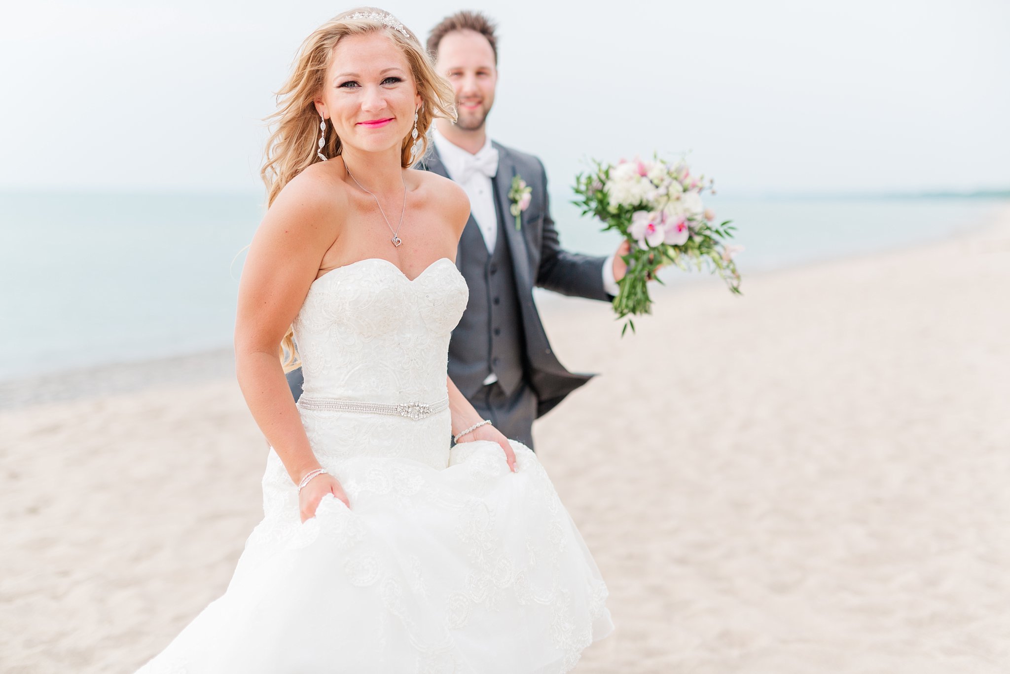 a bride and groom walk across a beach at oakwood resort. the bride is smiling at the camera and the groom is out of focus behind her. used in a blog post about london ontario wedding photographers
