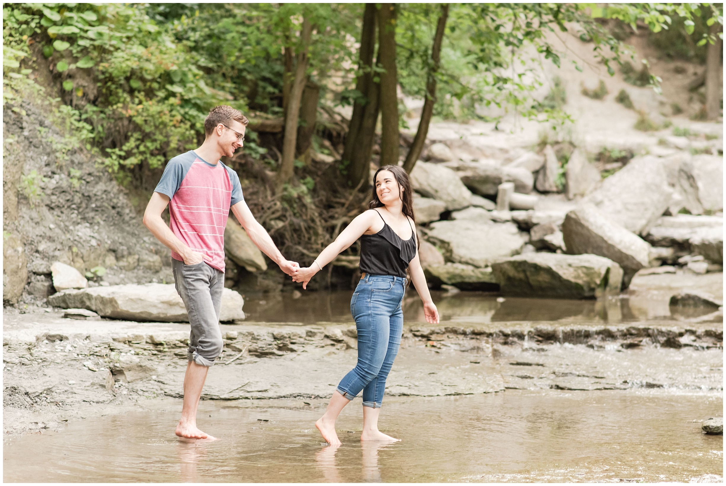 a woman wearing a black tank top and blue jeans leads a man in a pink shirt and grey shorts. they're walking through a shallow pond with trees and rocks in the background. photo is used in a blog post about social media for photographers