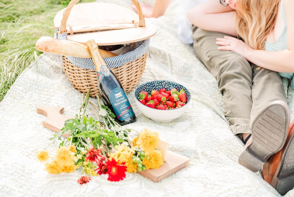 london ontario engagement photographer - photo of a bottle of champagne, some flowers, and a bowl of strawberries beside a picnic basket on top of a white blanket