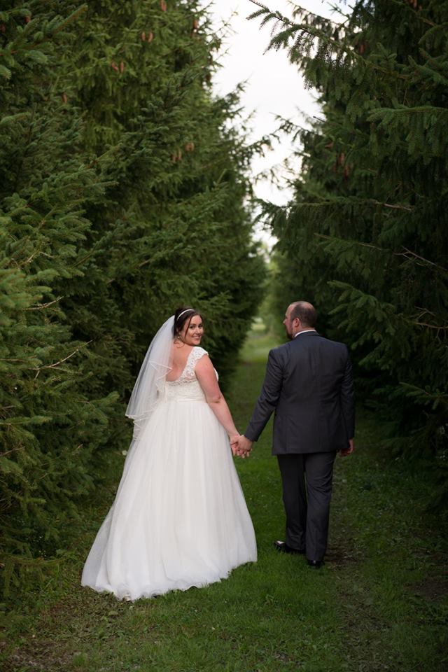 A bride and groom hold hands as they walk through a row of trees at Bellamere Winery. The groom is looking at the bride and the bride is looking back, smiling at the camera.