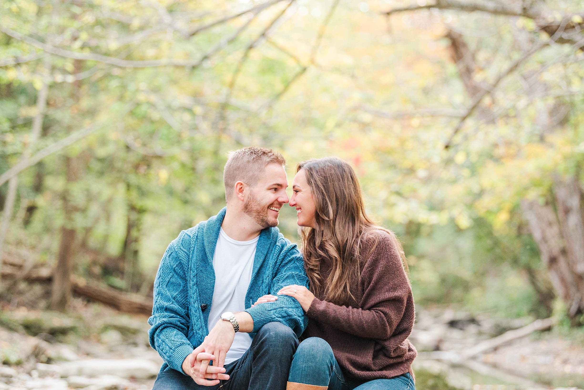 A man and woman smile at one another as they sit on the ground in front of a creek and full green and yellow fall trees during their Kelowna Wedding Photographer engagement session