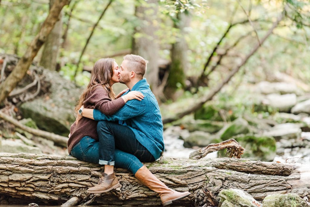 A couple sits facing one another on a fallen tree and they are kissing