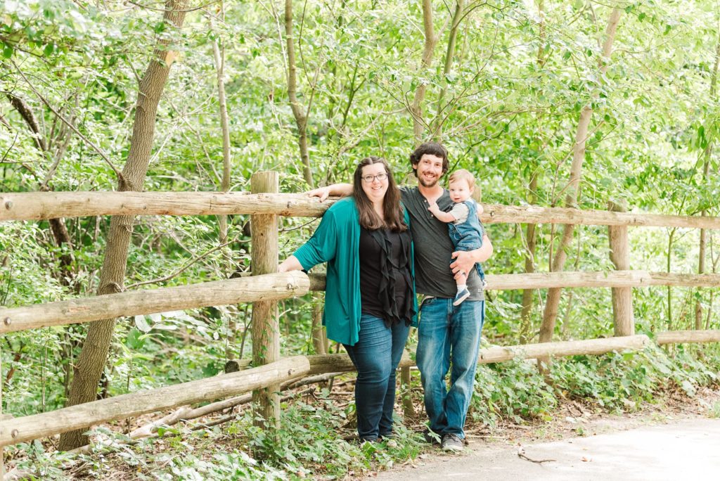 a family of three stands together beside a wooden fence
