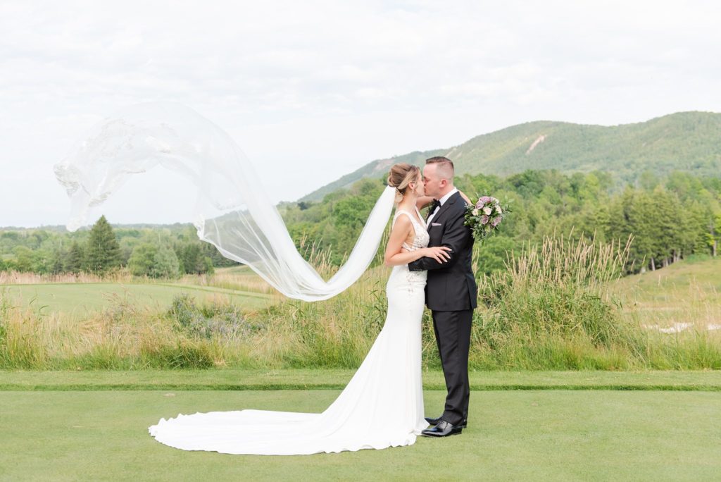 a bride's veil flies in the air as she and the groom kiss at blue mountain in collingwood ontario. kelowna wedding photography by life is beautiful photography