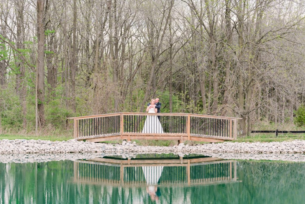 a bride and groom stand on a bridge, their reflection visible in the water below them