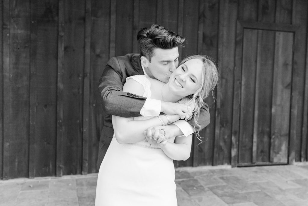 the groom wraps his arms around his bride and kisses her on the cheek