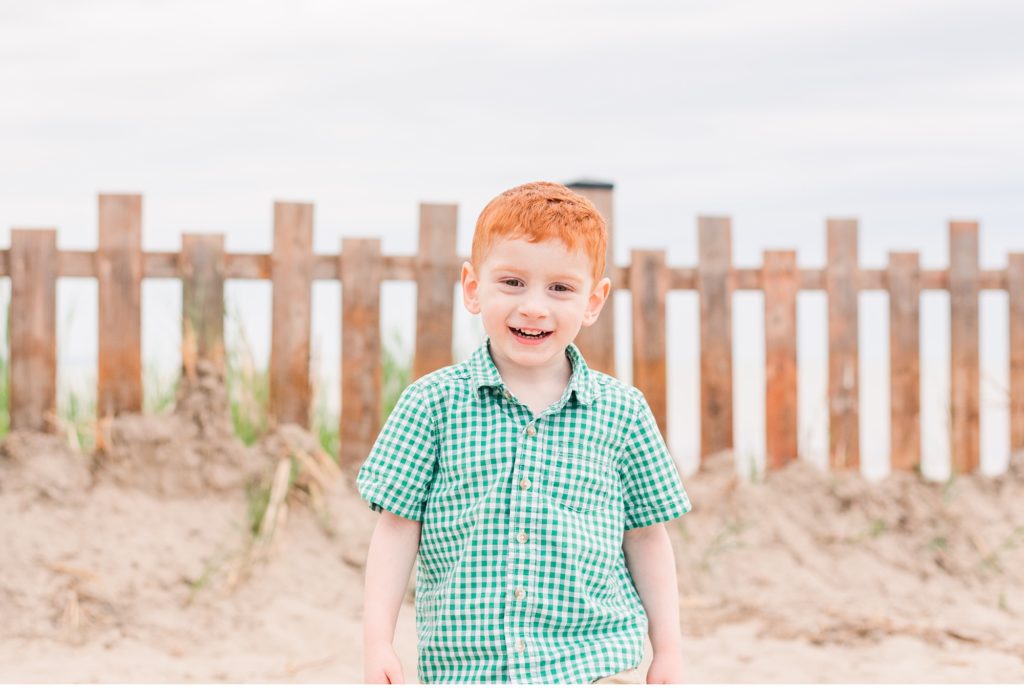 a little boy with red hair and a green plaid shirt smiles at the camera. a fence and sand are visible behind him. family photography in london ontario.