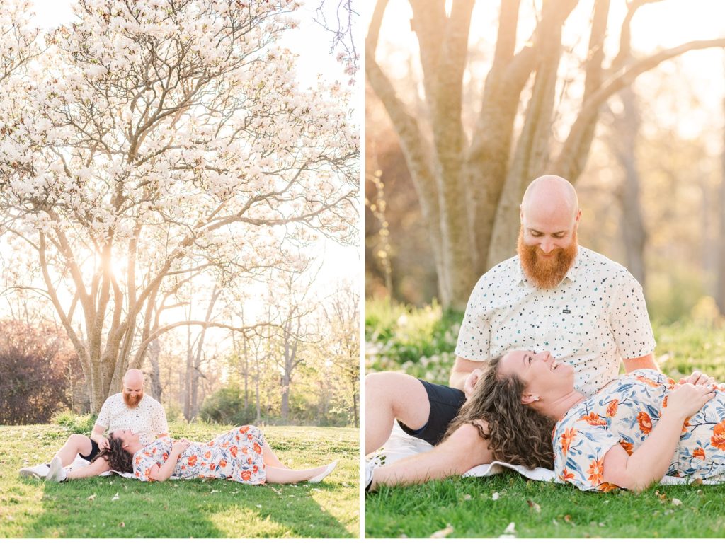 A man sits under a magnolia tree and his fiance lays down with her head in his lap during their engagement session with cherry blossoms in ontario