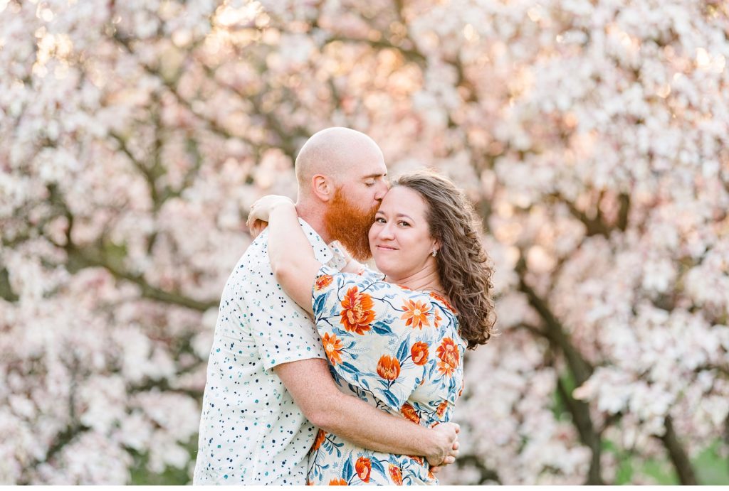 a man kisses a woman on her temple while she smiles at the camera during their engagement session with cherry blossoms in ontario